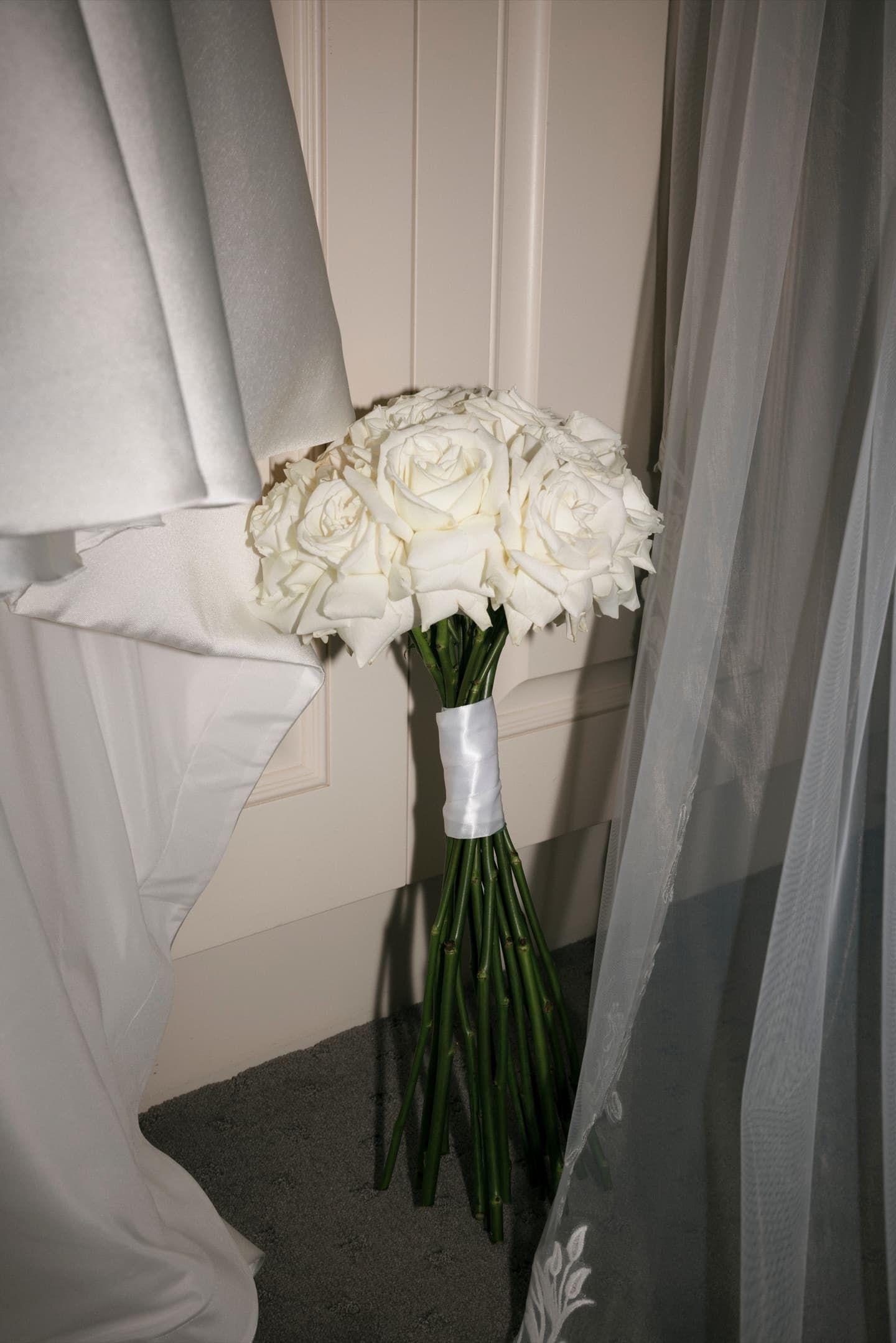 White rose bridal bouquet standing on satin-wrapped stems beside the gown and veil, designed by La Fleur Botanica