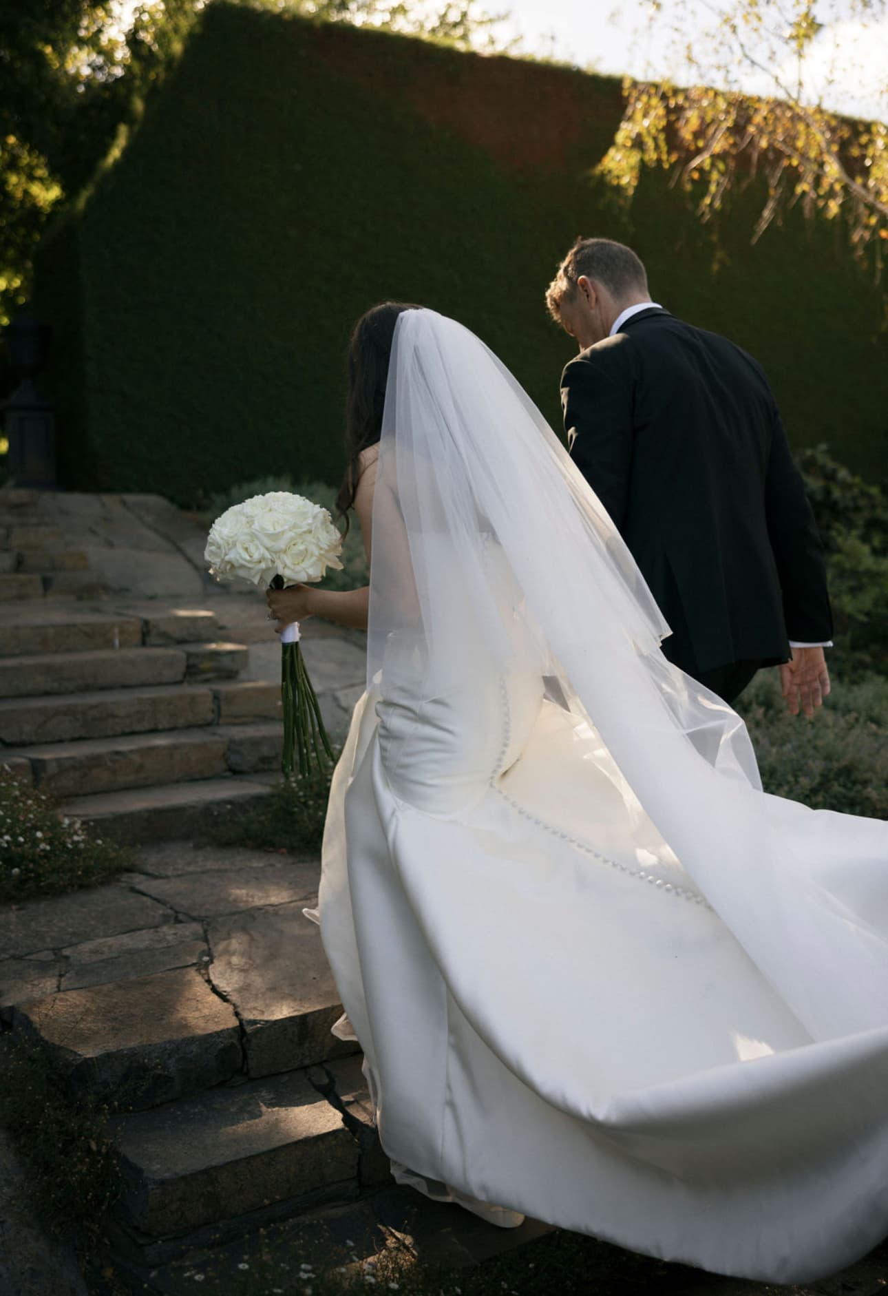 Bride and groom climbing stone garden steps at golden hour, florals by Hayley at La Fleur Botanica