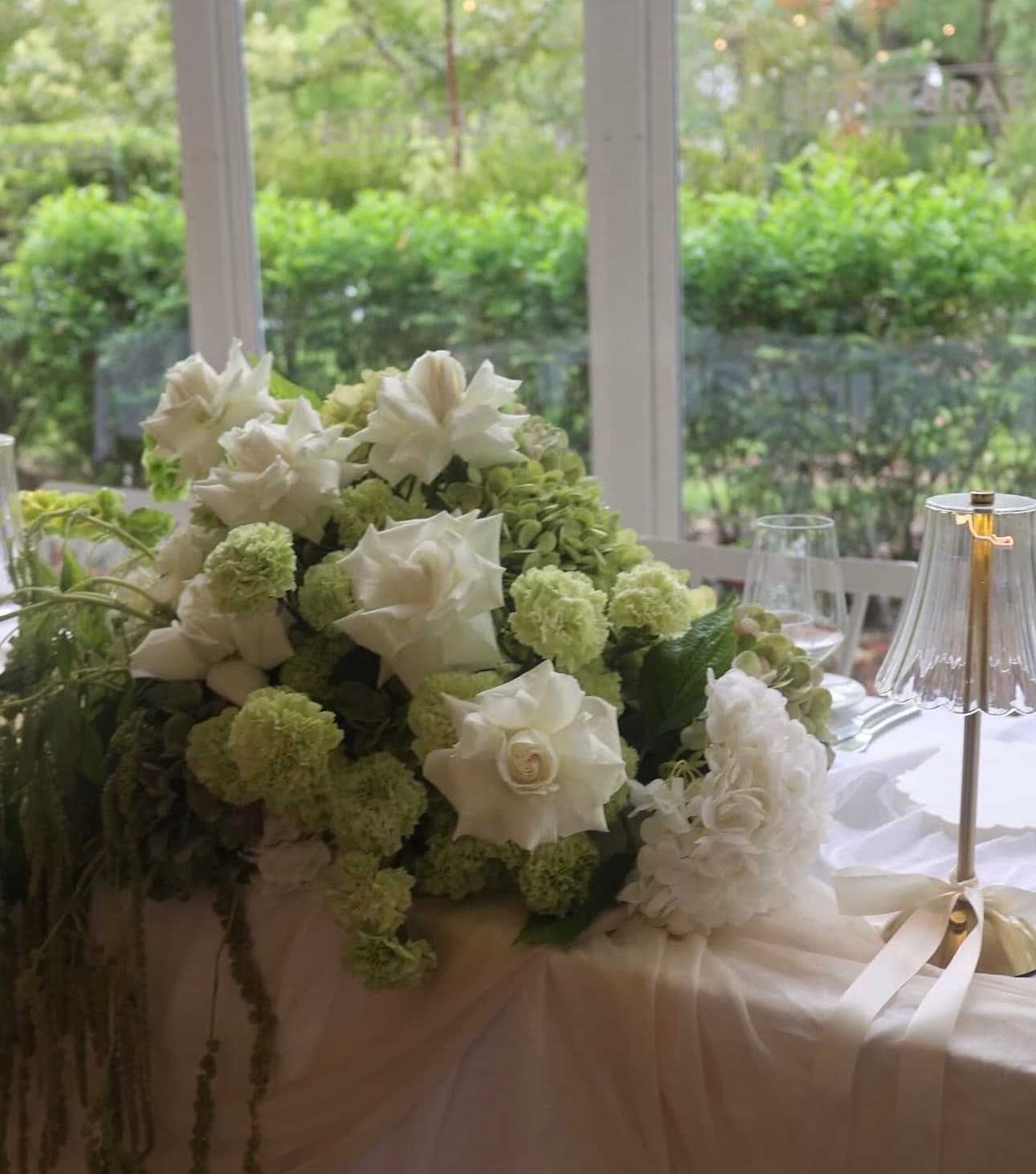 Long-table arrangement of white roses, green carnations and hydrangea, conservatory dining setting with crystal lamp, designed by La Fleur Botanica